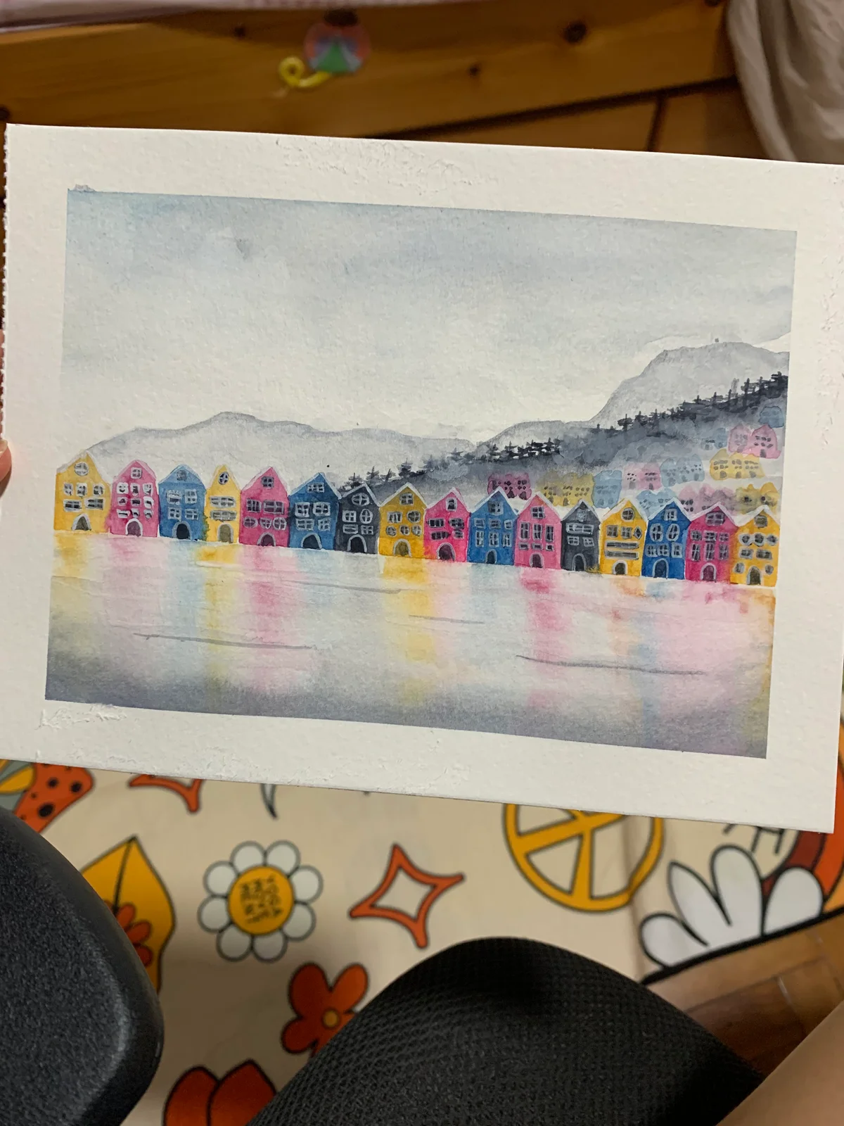 Painting of colourful waterfront houses reflected in water with hills in the background.