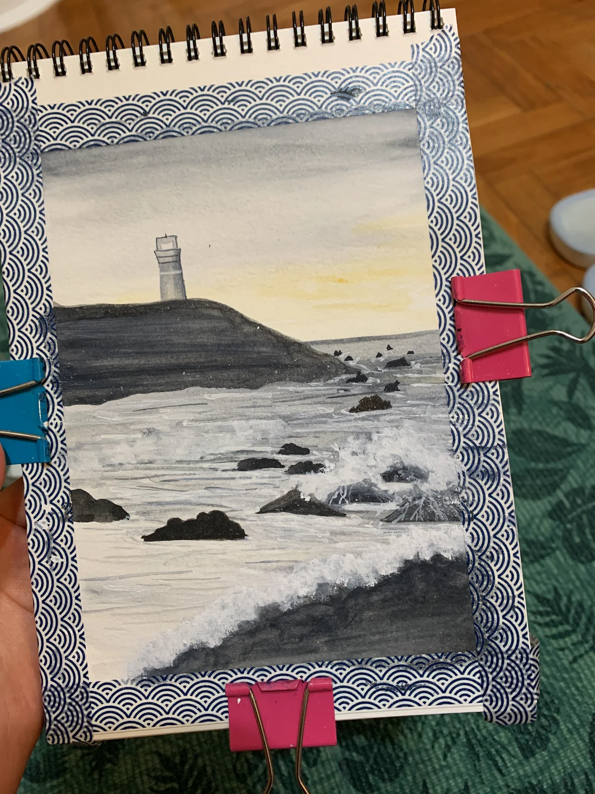 Sketchbook painting of waves and dark rocks below a coastal tower on a headland.